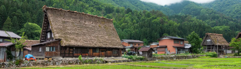 Traditional Japanese thatched-roof houses are surrounded by lush green mountains and rice fields in a rural village, with cloudy skies above.
