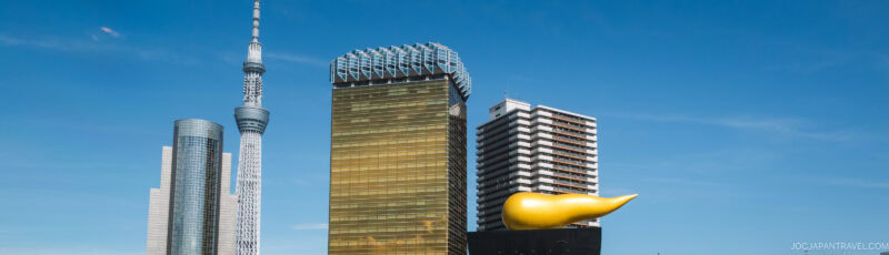 Tokyo skyline featuring the Tokyo Skytree, a modern glass building, the gold Asahi Beer Hall with its distinctive golden flame-shaped sculpture, and another high-rise against a clear blue sky.