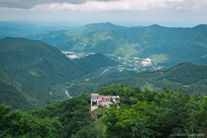 A scenic view of the lush green mountains of Nikko Akechidaira Plateau, featuring a small building and cable car station in the foreground, overlooking a valley with winding roads and distant villages under a cloudy sky.