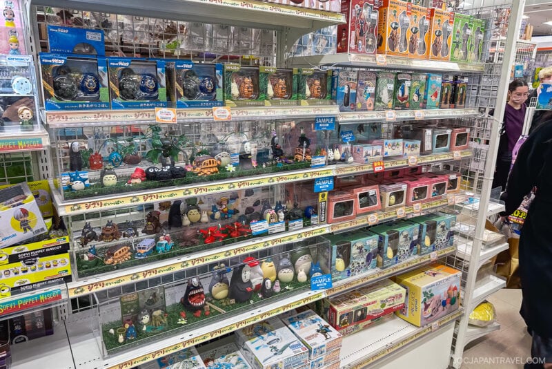 A store shelf in Akihabara Hobby Stores displays various Studio Ghibli merchandise, including figurines, music boxes, and themed collectibles from Totoro, Spirited Away, and other films. Shoppers are visible in the background.