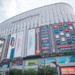 Large electronics store building near Akihabara Station in Tokyo, with multiple colorful signs and advertisements for gadgets, cameras, and phones. The vibrant, modern building is bustling, and most of the signage is in Japanese.