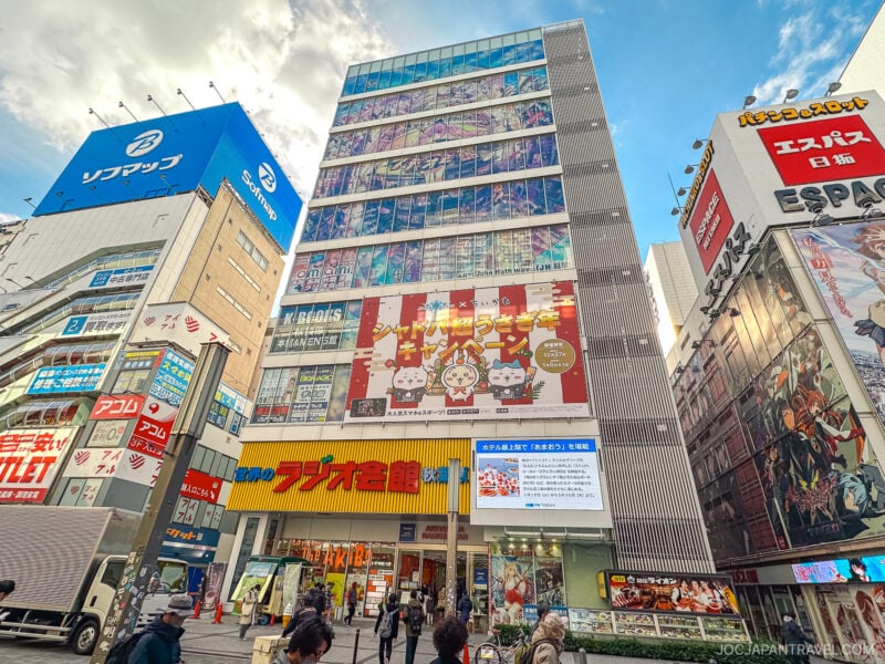 A busy street scene near Akihabara Station in Tokyo, with tall buildings covered in colorful anime and electronic store advertisements, people walking, and a partly cloudy sky overhead.