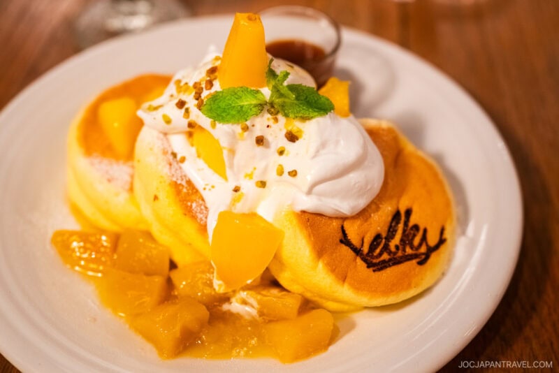 Fluffy Japanese pancakes at Amerikamura Elk Osaka, topped with whipped cream, diced mango, chopped nuts, and a mint sprig, served with syrup on a white plate. The word Lovely is written on one pancake.