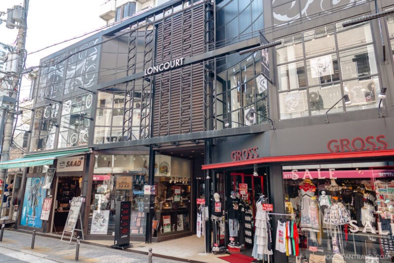 Street view of a modern building in Amerikamura with glass storefronts, featuring the LONCOURT entrance in the center, GROSS clothing store with red accents on the right, and another shop offering various items on the left.
