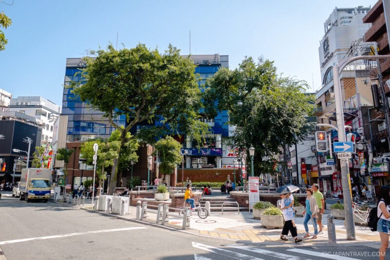 A lively Amerikamura city street scene in Japan with people walking, trees lining the sidewalk, modern buildings in the background, and a small plaza area in the center. Bright, sunny weather.