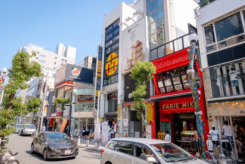A busy street in a Japanese city’s Amerikamura district with colorful storefronts like Paris Miki and Burger King, cars passing by, trees along the sidewalk, and pedestrians strolling in bright daylight.
