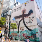 Amerikamura (アメリカ村) – Osaka’s Hub for Fashion, Art, and Youth Culture A large mural in Amerikamura shows a black bird with flaming wings and the words PEACE ON EARTH. Below, colorful graffiti covers the lower wall as people stroll past lively shops and street stalls under a clear sky.