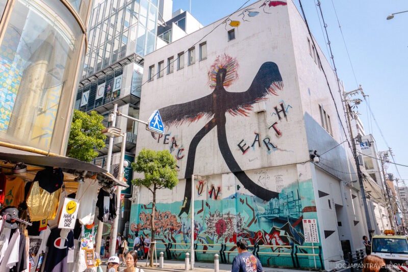 A large mural in Amerikamura shows a black bird with flaming wings and the words PEACE ON EARTH. Below, colorful graffiti covers the lower wall as people stroll past lively shops and street stalls under a clear sky.