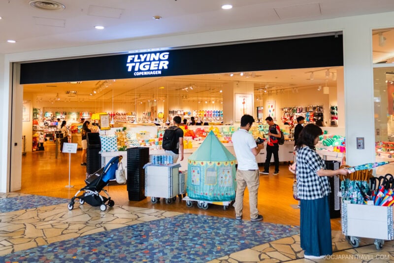 A brightly lit Flying Tiger Copenhagen store in Aqua City Odaiba with shoppers browsing colorful items, toys, and home goods. A stroller, a play tent, and baskets are visible near the entrance.