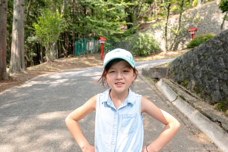 A young girl in a light blue sleeveless shirt and cap stands on a sunlit path at Arakurayama Sengen Park, smiling with her hands on her hips. Trees and stone walls line the background.