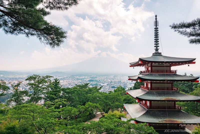 A traditional five-story pagoda stands among green trees in Arakurayama Sengen Park, with a distant city view and hazy Mount Fuji partially visible through clouds in the background.