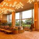 Spacious, elegant Arima Grand Hotel lobby with modern chandeliers, floor-to-ceiling windows, lush indoor plants, and comfortable seating. Large windows showcase a green garden outside. Warm lighting creates a welcoming atmosphere.