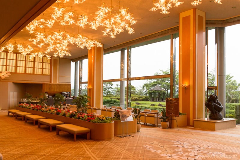 Spacious, elegant Arima Grand Hotel lobby with modern chandeliers, floor-to-ceiling windows, lush indoor plants, and comfortable seating. Large windows showcase a green garden outside. Warm lighting creates a welcoming atmosphere.