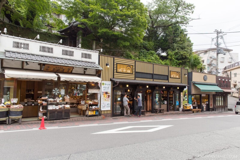 A street in Arima Onsen, Japan, with traditional-style shops and cafes, some displaying goods outside. Trees and greenery are visible behind the buildings and a few people are standing near the entrances.