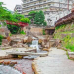 Arima Onsen (有馬温泉) – One of Japan’s Three Great Hot Springs A stone walkway crosses a stream with small waterfalls in an urban park at Arima Onsen, surrounded by greenery and buildings. A red bridge and pedestrians are visible in the background.