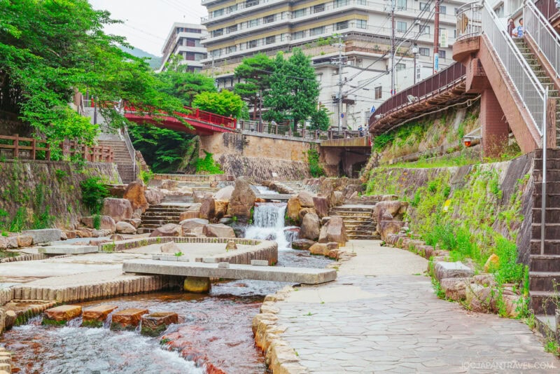 A stone walkway crosses a stream with small waterfalls in an urban park at Arima Onsen, surrounded by greenery and buildings. A red bridge and pedestrians are visible in the background.