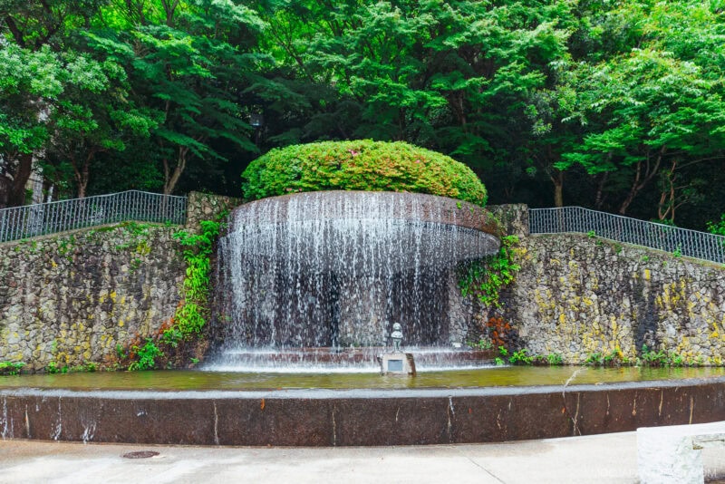 A circular stone fountain with water cascading down its sides, inspired by Arima Onsen, surrounded by lush green trees and plants, and a small statue at the center of the water basin in front.