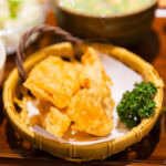 A close-up of assorted tempura pieces, including Beppu Chicken Tempura Toriten, in a bamboo basket, served with parsley garnish, alongside a bowl of rice and other side dishes on a wooden tray.