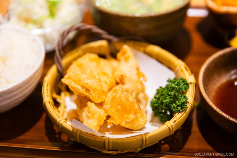 A close-up of assorted tempura pieces, including Beppu Chicken Tempura Toriten, in a bamboo basket, served with parsley garnish, alongside a bowl of rice and other side dishes on a wooden tray.