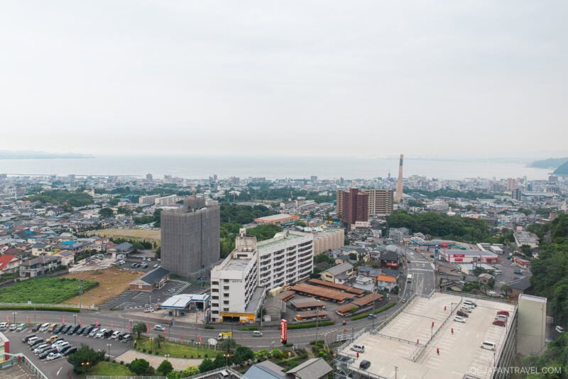 Aerial view of a coastal city featured in the Beppu Onsen Travel Guide, with clustered buildings, roads, green patches, and a distant body of water under a cloudy sky. Cars are parked in lots as the city stretches toward the shoreline.