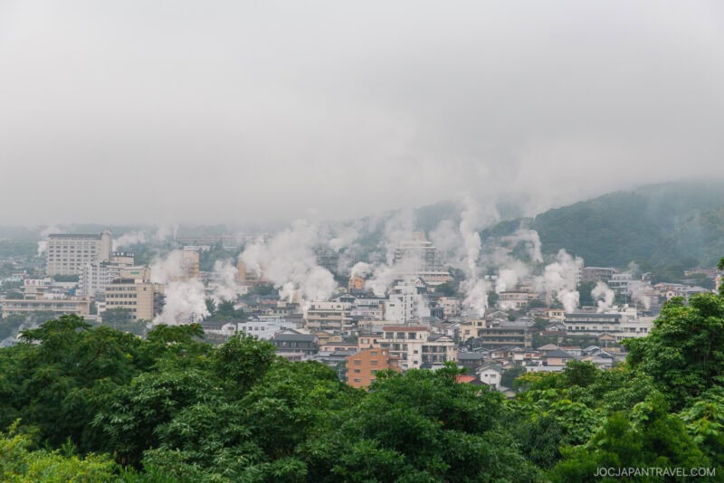 A cityscape with numerous buildings emitting white steam, surrounded by green hills and partially obscured by fog or low clouds—a scene straight from a Beppu Onsen Travel Guide. Dense greenery fills the foreground.