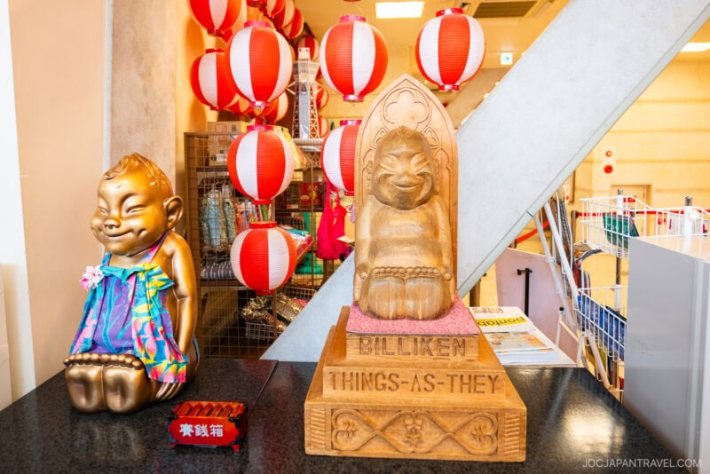 Two Billiken Osaka statues&mdash;one gold and wrapped in colorful cloth, the other wooden and inscribed with BILLIKEN THINGS-AS-THEY&mdash;stand on a counter, with red and white paper lanterns hanging in the background.