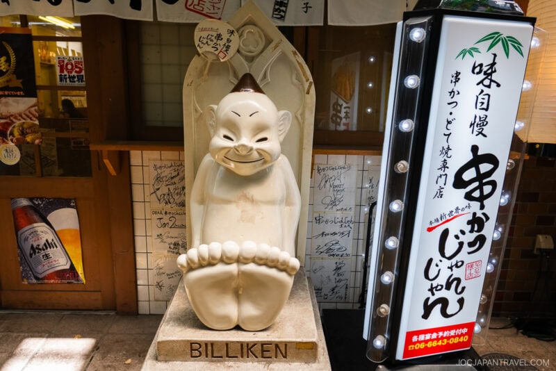 A statue of Billiken Osaka, the chubby figure with pointed ears and a wide grin, sits displayed outside a Japanese restaurant, with signed papers on the wall and a bright vertical sign adorned with Japanese text beside it.