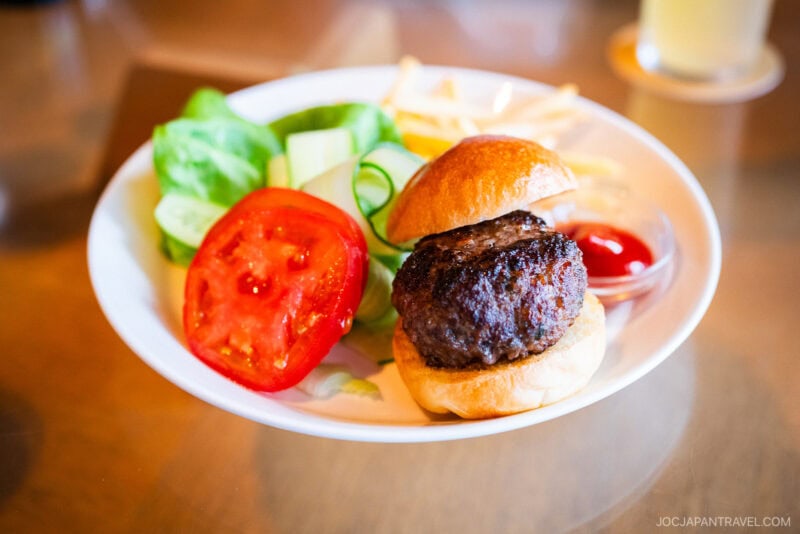 A plate at Bills Odaiba at DECKS features a hamburger on a bun with lettuce, tomato slices, cucumber, a small serving of fries, and a dollop of ketchup, all presented on a wooden table.