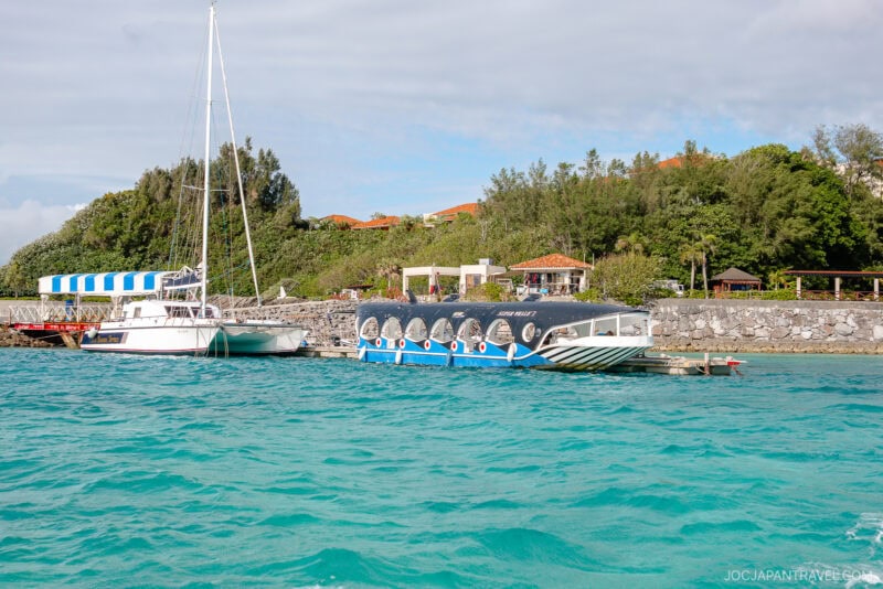 A glass-bottom boat and a sailboat are docked by a rocky shoreline with turquoise water at Busena Marine Park. Green trees and red-tiled roof buildings stand in the background beneath a partly cloudy sky.
