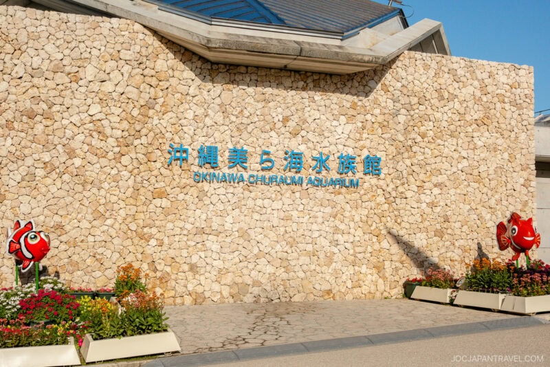 The entrance of Churaumi Aquarium features a stone wall with blue signage in Japanese and English, flanked by flowerbeds and playful cartoon clownfish statues on each side.