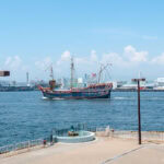 Cruise Ship Santa Maria – Set Sail Around Osaka Bay on a Replica Galleon A large, colorful ship styled like the Osaka Cruise Ship Santa Maria sails in a harbor near an industrial cityscape, with cranes and warehouses in the background and a clear blue sky overhead.