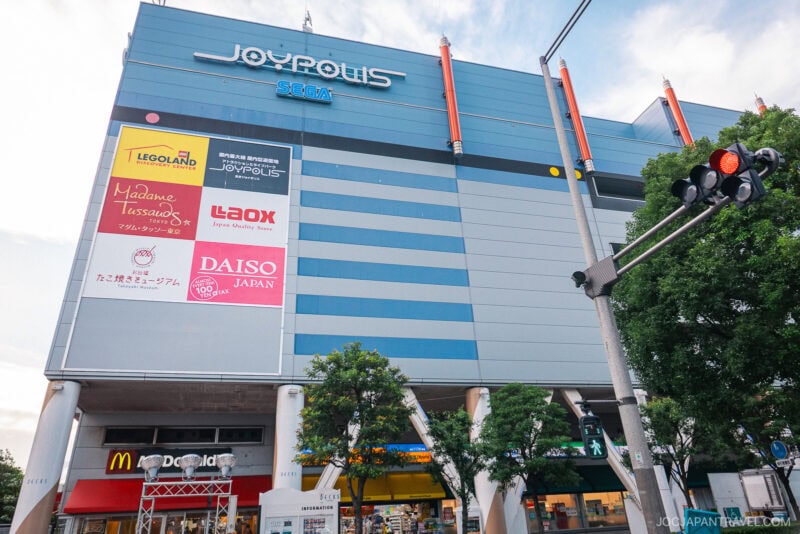 A large blue Joypolis SEGA building at DECKS Tokyo Beach features store signs like LEGOLAND, Madame Tussauds, Daiso Japan, and Laox. Trees line the sidewalk, and a McDonalds is visible at street level.