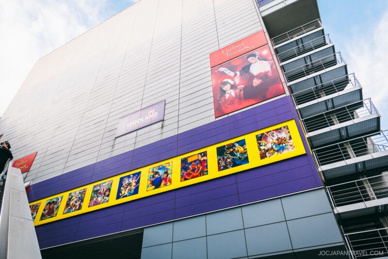 A modern building at DECKS Tokyo Beach showcases colorful LEGO-themed murals and a LEGOLAND sign, with bright artwork and posters, including one for Harry Potter and the Chamber of Secrets. Metal stairs are visible on the right side.