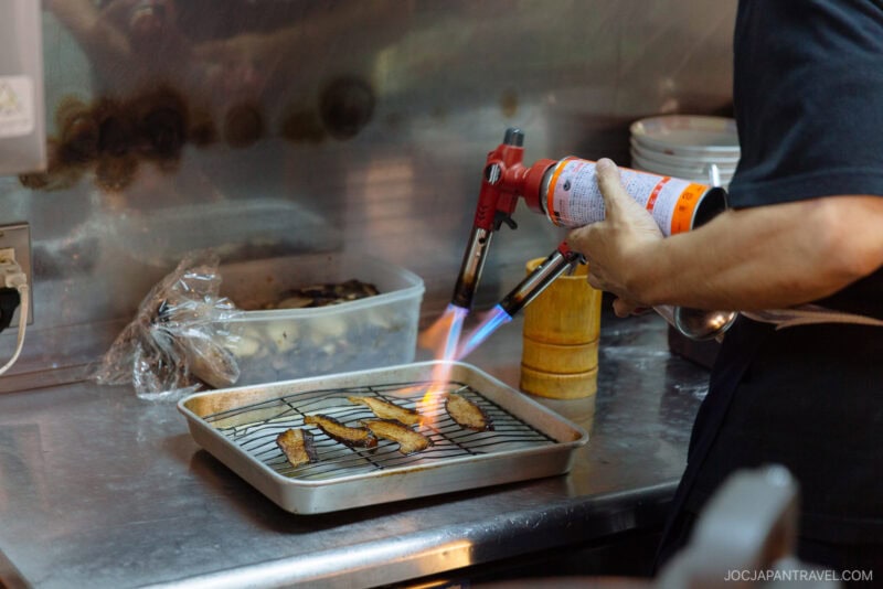 A person uses a handheld blowtorch to sear pieces of fish or meat on a wire rack over a tray in a kitchen at Daruma Ramen Fukuoka. Nearby, there is a plastic container with food and other kitchen items.