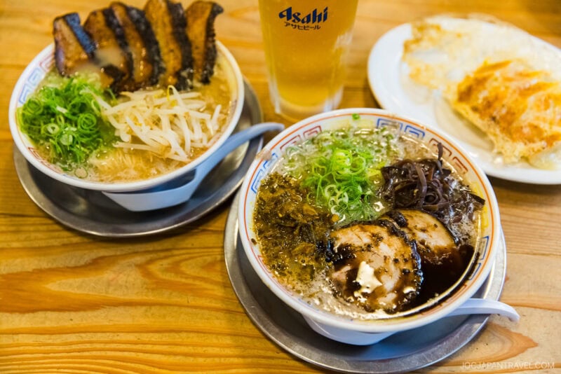 Two bowls of Daruma Ramen Fukuoka with sliced pork, green onions, and various toppings sit on a wooden table, accompanied by a plate of gyoza and a glass of Asahi beer in the background.
