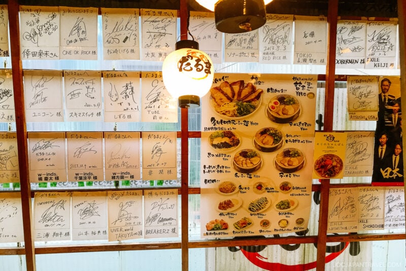 Wall decorated with autographed papers, a round white lantern with Japanese text, and a menu featuring various Japanese dishes and drinks. The setting appears to be inside Daruma Ramen Fukuoka, a cozy Japanese restaurant.