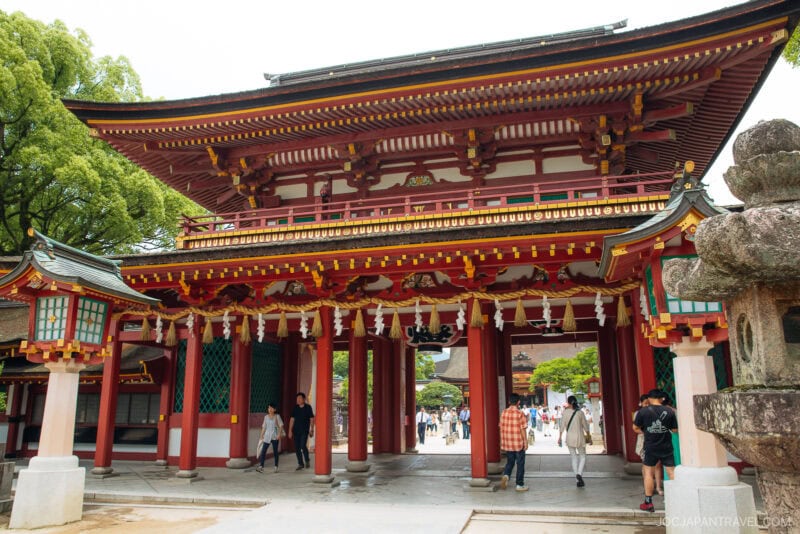 A large, ornate red and white wooden gate with traditional Japanese architectural features stands at the entrance of Dazaifu Tenmangu Shrine, as visitors walk through and around it on a sunny day.
