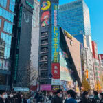 Akihabara Don Quijote (ドン・キホーテ) – Tokyo’s Wildest Discount Store A busy street in Tokyo’s Akihabara with crowds of people, many wearing masks, walking among tall buildings covered in colorful signs and ads, including a large Don Quijote store with vibrant anime-themed signage.