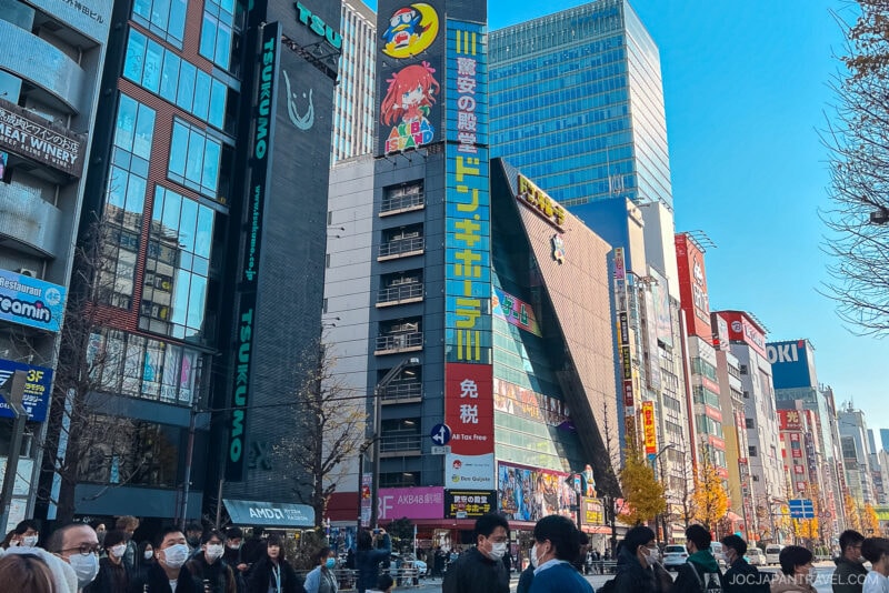 A busy street in Tokyo&rsquo;s Akihabara with crowds of people, many wearing masks, walking among tall buildings covered in colorful signs and ads, including a large Don Quijote store with vibrant anime-themed signage.