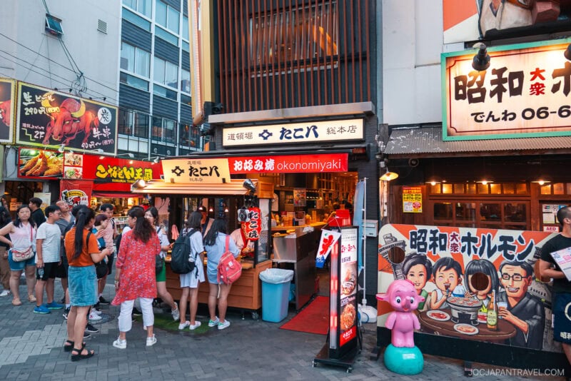 A group of people wait outside colorful Japanese restaurants with bright signs, including one for okonomiyaki and Dotonbori Takoyaki. Neon lights and illustrated menus decorate the scene, and a pink pig statue stands near the entrance.