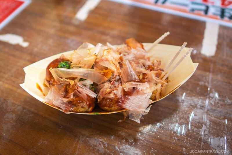 A bamboo tray filled with Dotonbori Takoyaki, Japanese octopus balls topped with bonito flakes, sauce, and herbs, sits on a wooden table with toothpicks for eating.