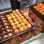 Rows of takoyaki (Japanese octopus balls) cooking in metal molds, some already golden and garnished. A person’s hand is partially visible, turning the Dotonbori Takoyaki on one side of the grill.