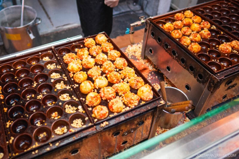 Rows of takoyaki (Japanese octopus balls) cooking in metal molds, some already golden and garnished. A person&rsquo;s hand is partially visible, turning the Dotonbori Takoyaki on one side of the grill.