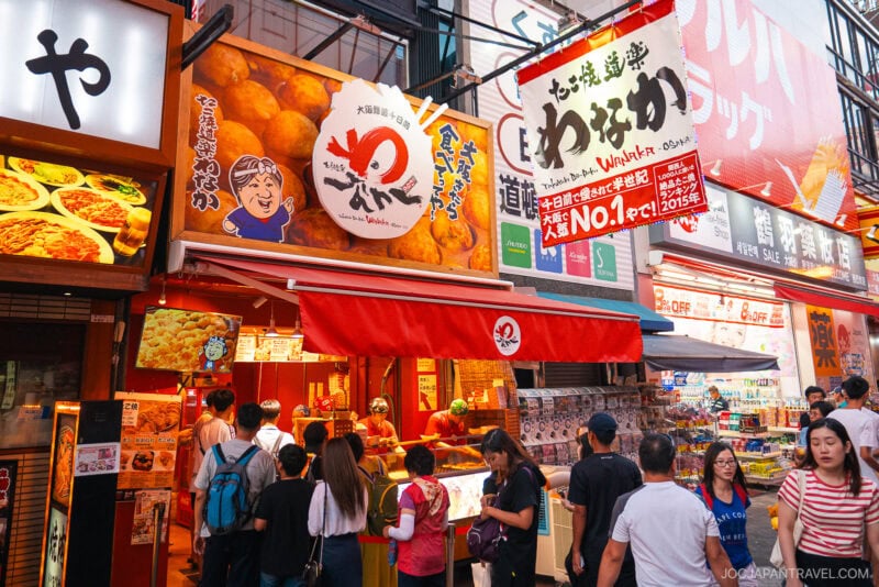 A busy street scene in Japan shows people gathered outside a bright red Dotonbori Takoyaki shop decorated with large signs, cartoon graphics, and colorful menus under the glow of market lights.