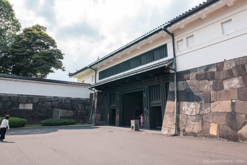 A wide stone gate with high white walls marks the entrance to Tokyo’s Imperial Palace, near the East Gardens of the Imperial Palace, with a few people walking and trees in the background under a partly cloudy sky.