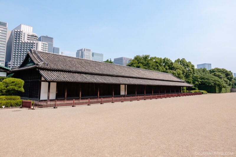 A traditional Japanese wooden building with a tiled roof stands in the East Gardens of the Imperial Palace, surrounded by trees and a wide gravel courtyard, with modern skyscrapers rising in the background under a clear blue sky.