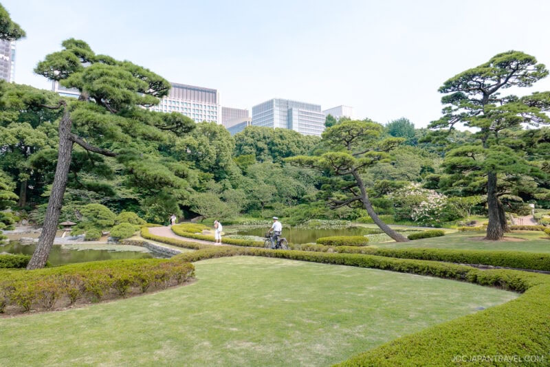 A serene Japanese garden inspired by the East Gardens of the Imperial Palace features carefully pruned trees, manicured lawns, winding gravel paths, and a pond. People stroll peacefully as modern city buildings rise in the background.