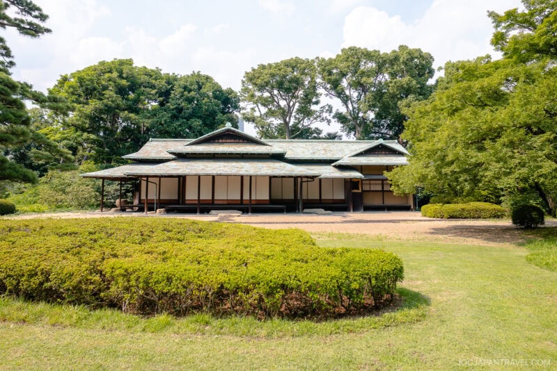 A traditional Japanese building with a green-tiled roof stands surrounded by manicured bushes, trees, and a well-kept lawn in the East Gardens of the Imperial Palace on a sunny day.