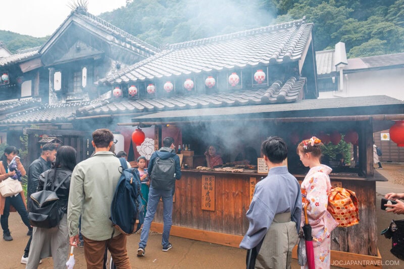 People gather at a traditional Japanese food stall in Edo Wonderland Nikko Edomura, surrounded by smoky air, wooden architecture, hanging lanterns, and lush greenery; one woman wears a pink floral kimono.