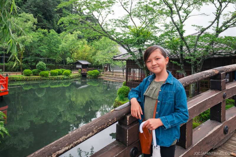 A young girl stands on a wooden bridge in a Japanese garden at Edo Wonderland Nikko Edomura, smiling at the camera. Behind her are lush green trees, manicured shrubs, a peaceful pond, and traditional wooden buildings.